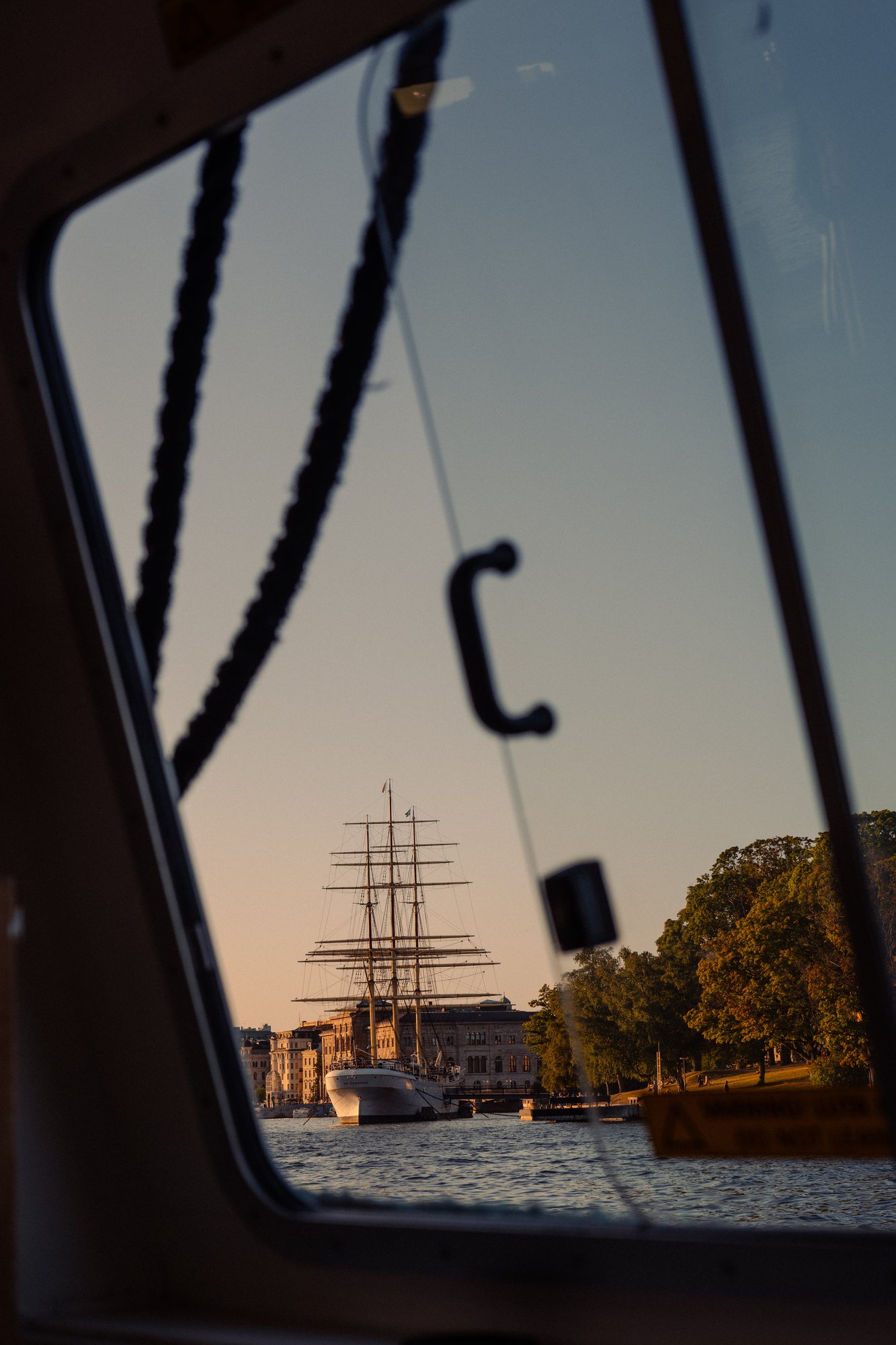 A tall ship seen through the window of a passing ferry at golden hour