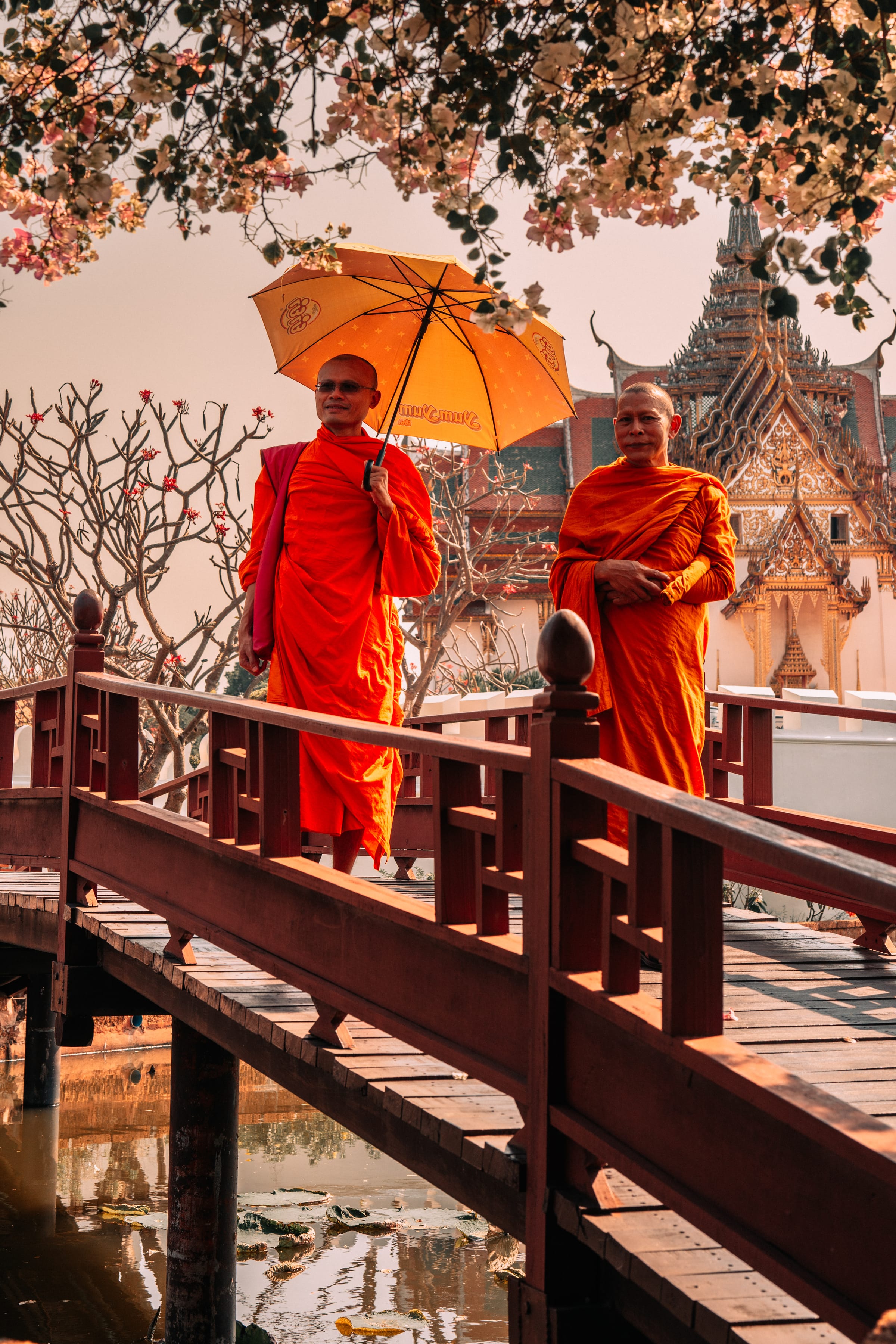 Two monks crossing a red wooden bridge under a single orange umbrella.