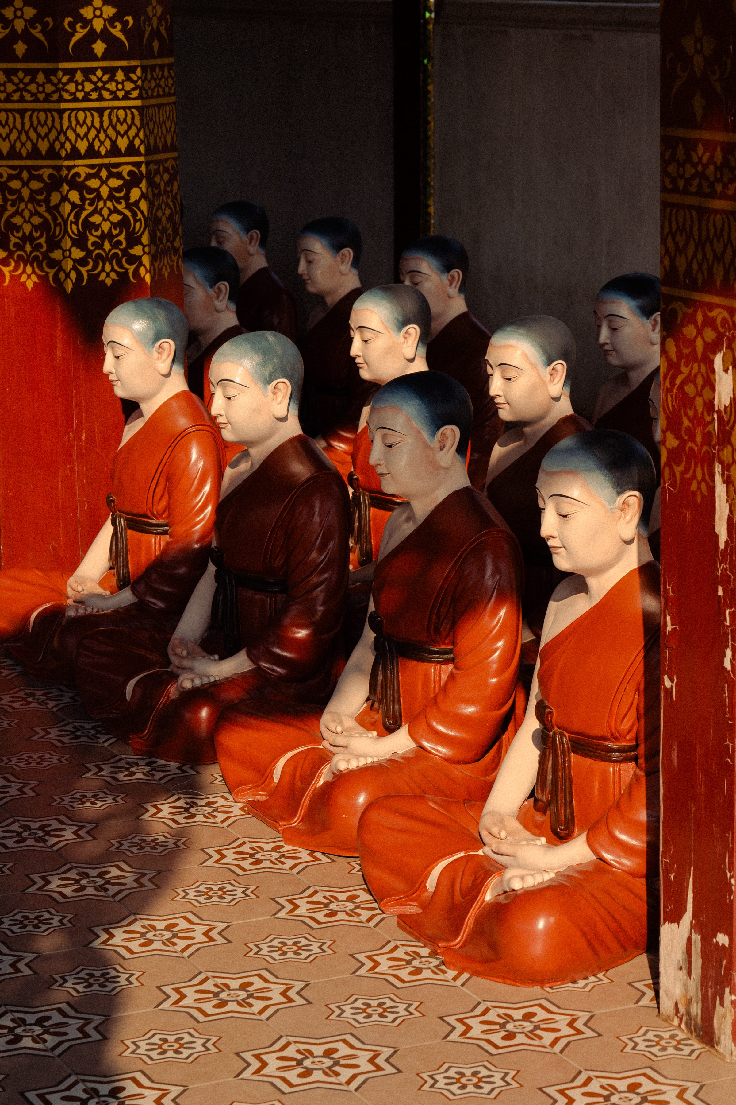 Rows of seated monk statues in orange robes, heads bowed and eyes lowered.