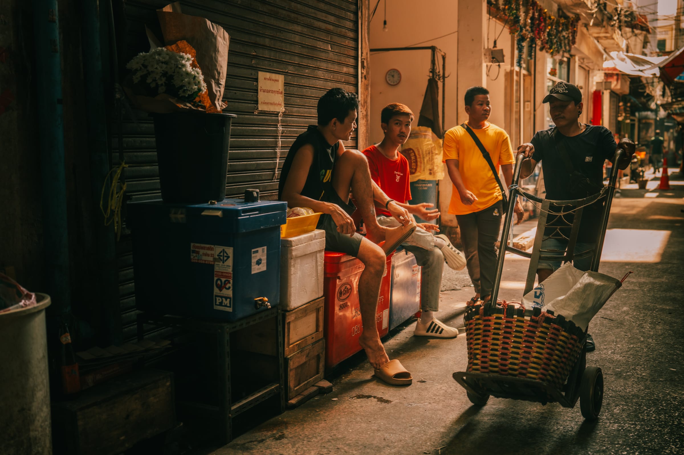 Four young men resting on crates in a narrow Chinatown alley, one with a hand trolley.