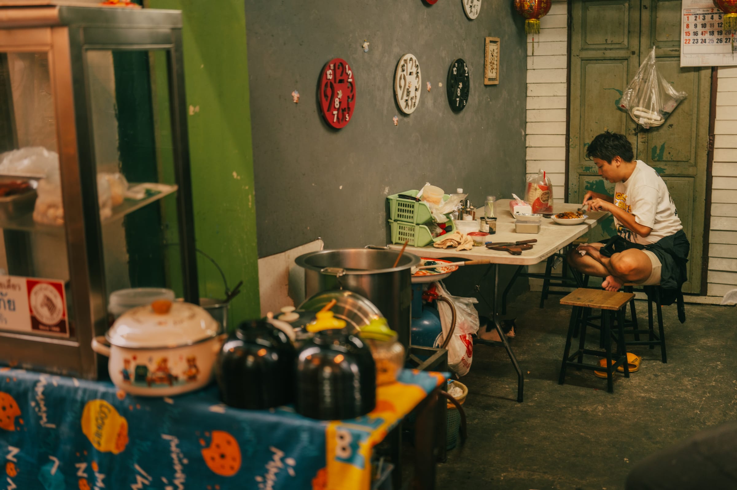 A man eating alone at a table beneath a wall of mismatched clocks.